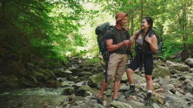 Gülümseyen çok uluslu sırt çantalı çift ormandaki dağ nehrinin yakınındaki haritada konuşuyorlar ve güzergahlarını kontrol ediyorlar.
