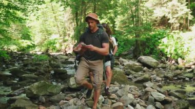 Yoğunlaştırılmış çokuluslu sırt çantalı arkadaşlar haritadaki güzergahı kontrol ediyorlar ve ormandaki dağ nehrini takip ediyorlar.
