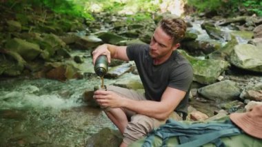 Kendine güvenen sarışın adam ormandaki dağ nehrinin yanındaki termostan çay içiyor.