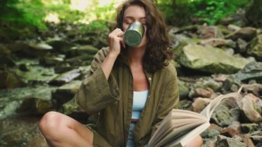 Güzel esmer turist kadın termostan çay içiyor ve ormanda dağ nehrinin yanında kitap okuyor.