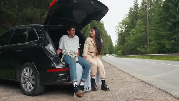 Stylish multinational couple talking in trunk of car in mountains