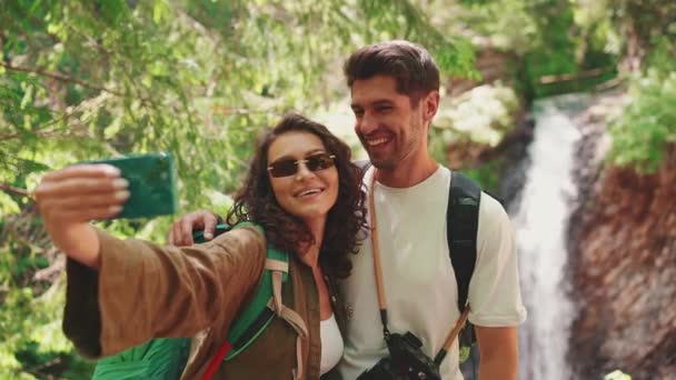 Uśmiechnięta Para Turystyczna Robi Sobie Selfie Przez Telefon Przeciwko Wodospadowi — Wideo stockowe