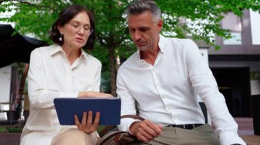 Konsantre olmuş yetişkin meslektaşlar, teras kafede iş yemeği yerken dijital tablet üzerindeki çalışmaları tartışırlar.