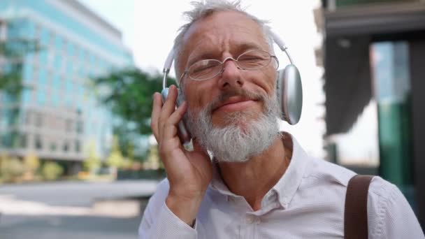 屋外で仕事をする途中で音楽を聴くスーツでリラックスした古いビジネスマン — ストック動画