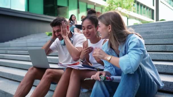 屋外の階段に座るラップトップの作業について話し合う多文化の若い学生を笑う — ストック動画