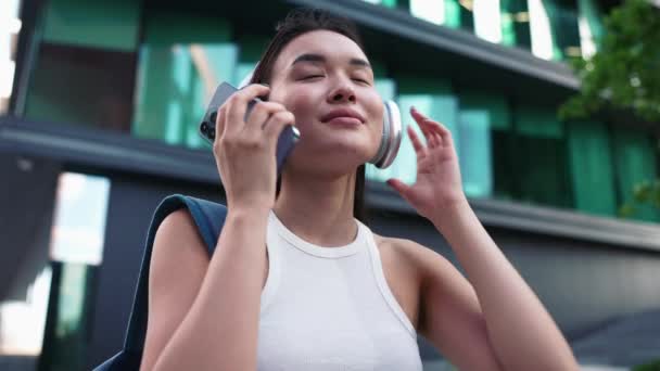 ハンサムな若い女性アジアの学生は 屋外でヘッドフォンで音楽を聞いています — ストック動画