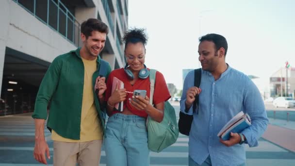 Hymyilevät Monirotuiset Opiskelijat Katselevat Matkapuhelinta Puhuvat Kävellessään Ulkona — kuvapankkivideo