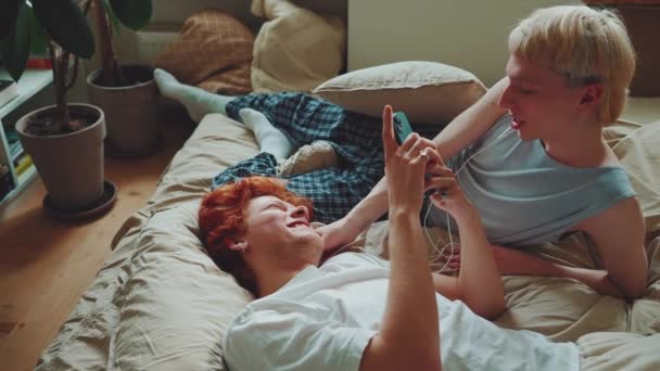 一对年轻有趣的同性恋夫妇 头发染得发亮 一边在家里用手机发短信 一边用耳机听音乐 — 图库视频影像
