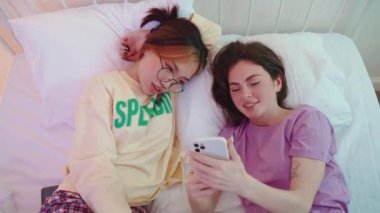 İki çok kültürlü, neşeli bayan arkadaş cep telefonuna bakıyor ve bir pijama partisinde yatakta konuşuyorlar.
