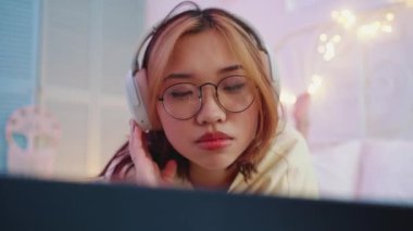 Gözlüklü gülümseyen Asyalı kadın kulaklıkla müzik dinliyor ve evdeki yatakta dizüstü bilgisayara bakıyor.