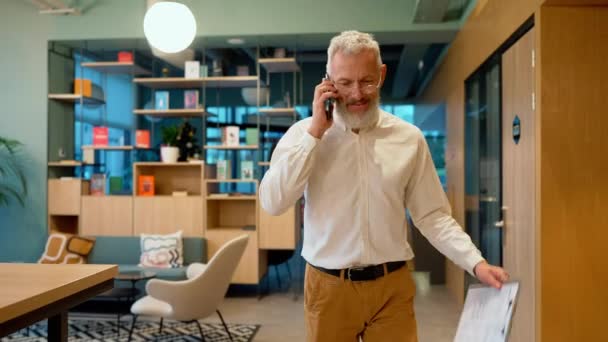 スマイリーグレーのビジネスマンは 電話で話し オフィスで歩いている間に紙を見ています — ストック動画