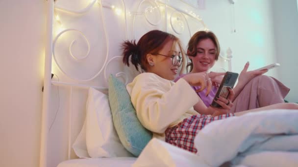 两个快乐的多文化女性朋友一边看手机 一边在床上通宵聊天 — 图库视频影像