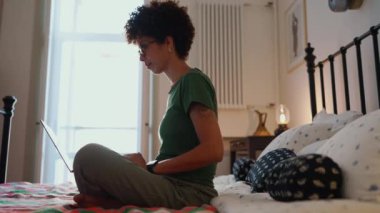 Gözlüklü, kıvırcık saçlı, özgüvenli Afrikalı kadın laptopta evden yatağa çalışıyor.