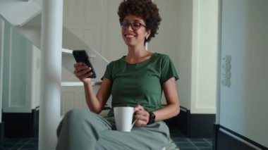 Gözlüklü, kıvırcık saçlı, telefonda kahve ile mesajlaşan pozitif Afrikalı bir kadın.