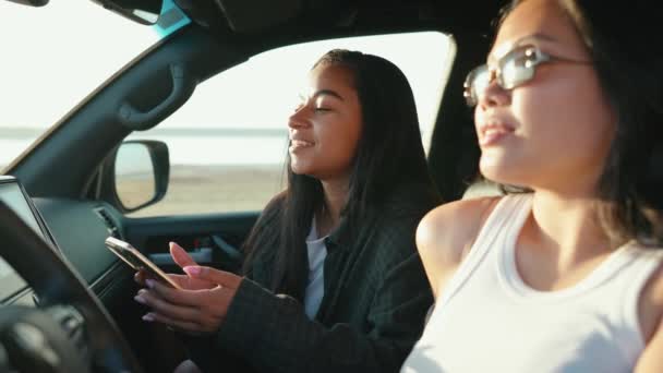 かわいい多文化の少女は 夏のエステリー近くの車の中で指を指し 携帯電話を見ています — ストック動画