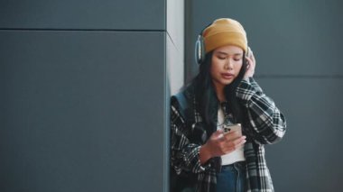 Rahatlamış Asyalı kadın, açık havada, siyah duvarın yanında kulaklıkla müzik dinliyor.