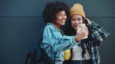 Çok kültürlü bayan arkadaşlar cep telefonlarında siyah duvarın yanında bir fincan kahve ile selfie çekiyorlar.