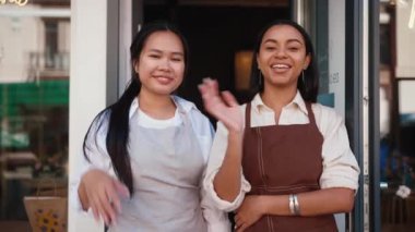 Positive multicultural waitresses in aprons waving hands at camera and welcoming guests at door of cafe