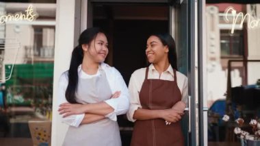 Happy multicultural waitresses in aprons welcoming guests at door of cafe