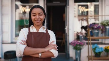 Laughing african waitress in apron looking at camera and welcoming guests at door of cafe