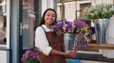 Handsome african waitress in apron welcoming guests with potted flowers at door of cafe