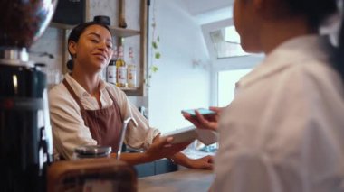 Smiling african female barista accepting bank card payments through terminal behind counter at cafe