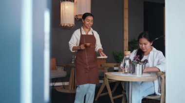 Cheerful african female barista bringing coffee to asian woman texting on cell phone in coffee shop