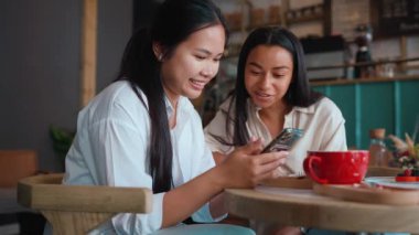Charming multicultural female friends looking at cell phone and talking with cup of tea at cafe