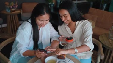 Laughing multicultural female friends looking at pictures on cell phone and eating cake at cafe