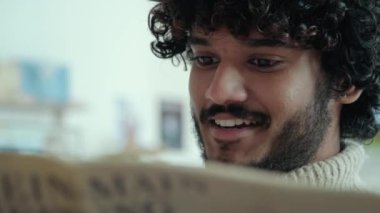 Smiling curly haired indian man in beige sweater reading newspaper sitting in cafe