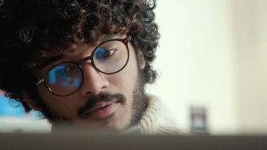 Pensive curly haired indian man in beige sweater and eyeglasses working on laptop in cafe