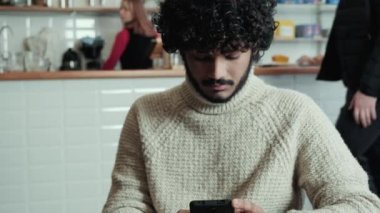 Concentrated curly haired indian man in beige sweater texting on mobile and looking around in cafe