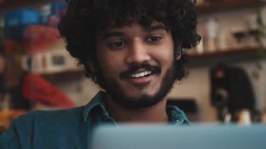 Charming curly haired indian man wearing jeans shirt talking by video call on laptop in cafe