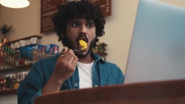 Surprised curly haired indian man wearing jeans shirt working on laptop and eating sponge cake in cafe
