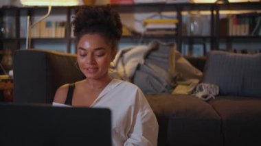 Gülen Afrikalı kadın dizüstü bilgisayarda çalışıyor ve akşam evde yerde otururken konuşuyor.