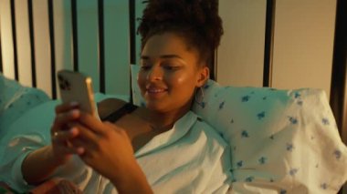 Neşeli Afrikalı kadın akşamları cep telefonuyla evde yatakta mesajlaşıyor.
