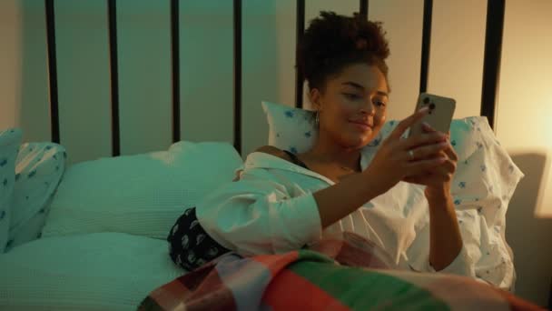夜に自宅でベッドでモバイルにタイピングするアフリカの女性を笑う — ストック動画