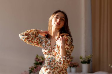 Pretty young woman in dress with flowers and dark hair is posing in sunset light in the white interior