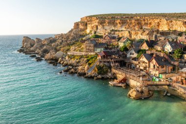 Malta, Il-Mellieha. Anchor Bay 'deki ünlü Temel Reis Köyü manzarası. Turkuaz deniz suları, mavi gökyüzü ve kayalıklarda gün batımı ışığı..