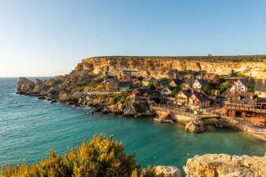 Malta, Il-Mellieha. Anchor Bay 'deki ünlü Temel Reis Köyü manzarası. Turkuaz deniz suları, mavi gökyüzü ve kayalıklarda gün batımı ışığı..