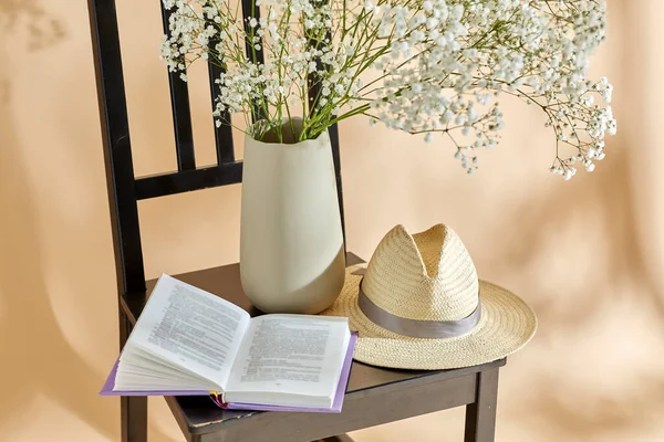 home decor and design concept - close up of gypsophila flowers in vase, book and straw hat on vintage chair over beige background
