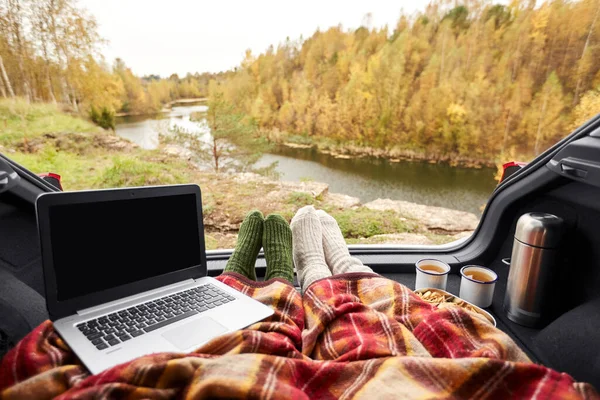 travel, tourism and camping concept - view to river from car trunk with couple feet in warm socks under blanket, two cups of tea with thermos, cookies and laptop computer