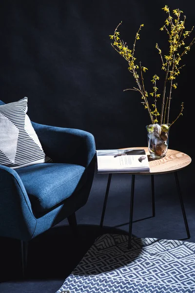 interior, holidays and home decor concept - close up of modern blue chair with pillow and easter eggs in vase with forsythia branches and magazine on table in dark room