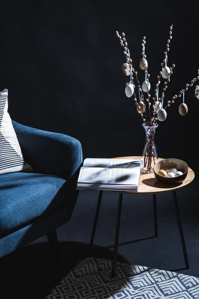 interior, holidays and home decor concept - close up of modern blue chair with pillow and easter eggs hanging on willow branches on table in dark room