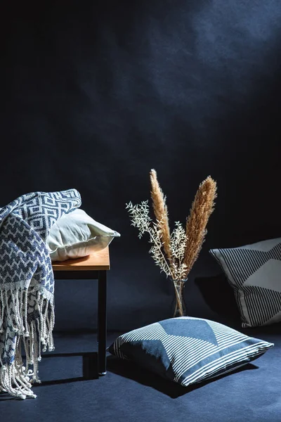 interior and home decor concept - pillows and blanket on bench and dry plants in vase on floor in dark room