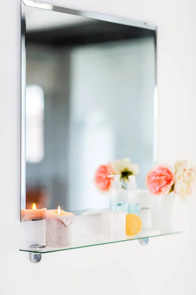 hygiene, beauty and daily routine concept - close up of lotion, sponge, cotton swabs with candle and flowers on mirror shelf in bathroom