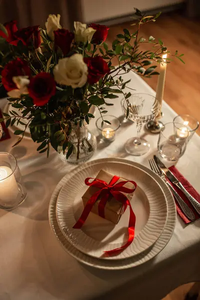holidays, dinner party and celebration concept - close up of festive table serving for two with gift box, flowers in vase and candles burning at home on valentines day