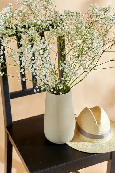 home decor and design concept - close up of gypsophila flowers in vase and straw hat on vintage chair over beige background