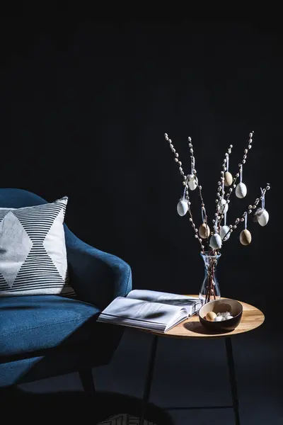 interior, holidays and home decor concept - close up of modern blue chair with pillow and easter eggs hanging on willow branches on table in dark room