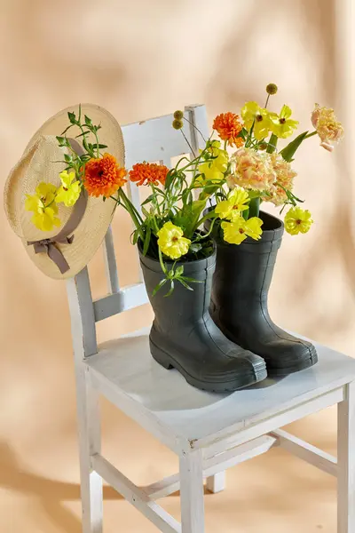 国际妇女日和花卉设计概念 米色背景的老式椅子上的橡胶靴和草帽花 免版税图库照片